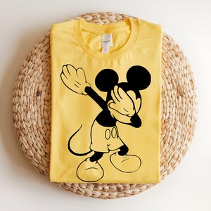 K&ouml;nnte beinhalten: Ein hellgelbes Kurzarm-T-Shirt mit einem schwarzen Grafikdruck von Mickey Mouse, der eine Dab-Pose ausf&uuml;hrt. Das Shirt hat einen Rundhalsausschnitt und ein wei&szlig;es Innenetikett, das am Kragen sichtbar ist, was ein klares Design zeigt.