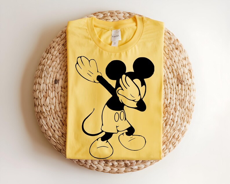 K&ouml;nnte beinhalten: Ein hellgelbes Kurzarm-T-Shirt mit einem schwarzen Grafikdruck von Mickey Mouse, der eine Dab-Pose ausf&uuml;hrt. Das Shirt hat einen Rundhalsausschnitt und ein wei&szlig;es Innenetikett, das am Kragen sichtbar ist, was ein klares Design zeigt.