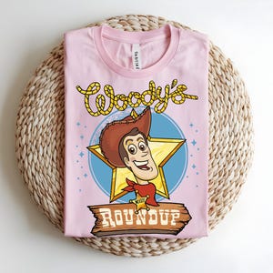 Puede incluir: Una camiseta rosa claro con una ilustraci&oacute;n de dibujos animados de Woody de Toy Story. El dise&ntilde;o incluye la cara de Woody, una estrella y las palabras "Woody's Roundup" en una fuente de estilo occidental. La camiseta est&aacute; doblada sobre una superficie tejida.
