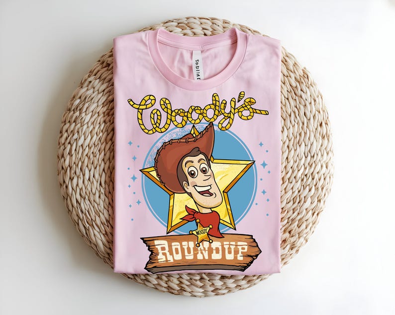 Puede incluir: Una camiseta rosa claro con una ilustraci&oacute;n de dibujos animados de Woody de Toy Story. El dise&ntilde;o incluye la cara de Woody, una estrella y las palabras "Woody's Roundup" en una fuente de estilo occidental. La camiseta est&aacute; doblada sobre una superficie tejida.
