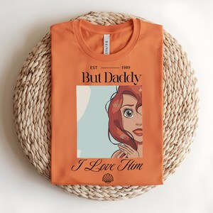 May include: An orange t-shirt featuring a graphic print of a red-haired character with the text "EST 1989", "But Daddy", and "I Love Him" in black lettering, along with a small black seashell icon centered at the bottom of the graphic design.