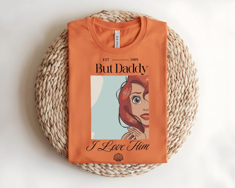 May include: An orange t-shirt featuring a graphic print of a red-haired character with the text "EST 1989", "But Daddy", and "I Love Him" in black lettering, along with a small black seashell icon centered at the bottom of the graphic design.