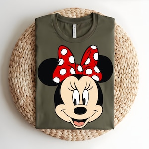 Puede incluir: Una camiseta de color verde oliva con un estampado gr&aacute;fico del rostro de Minnie Mouse. El personaje tiene orejas negras, cara de color crema y un gran lazo rojo con lunares blancos. La camiseta tiene cuello redondo y una etiqueta blanca.