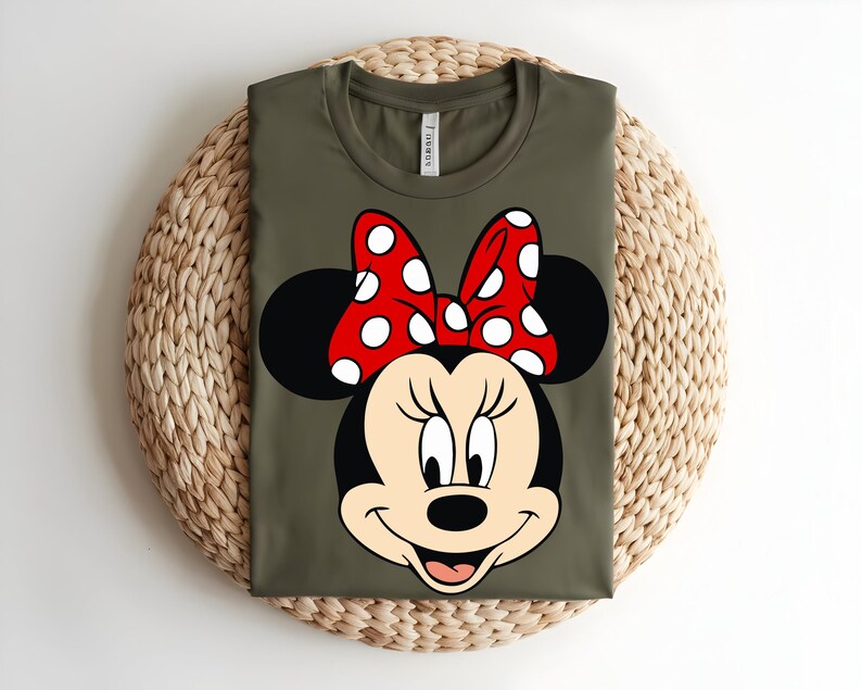 Puede incluir: Una camiseta de color verde oliva con un estampado gr&aacute;fico del rostro de Minnie Mouse. El personaje tiene orejas negras, cara de color crema y un gran lazo rojo con lunares blancos. La camiseta tiene cuello redondo y una etiqueta blanca.
