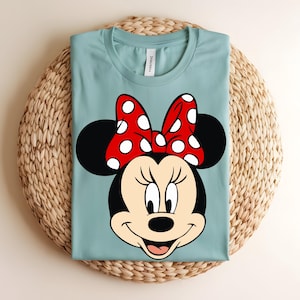 Puede incluir: Una camiseta de cuello redondo en color azul claro con un gr&aacute;fico grande del rostro de Minnie Mouse. El personaje tiene orejas negras, un lazo rojo con lunares blancos y una expresi&oacute;n sonriente. La camiseta tiene una etiqueta blanca en el cuello.
