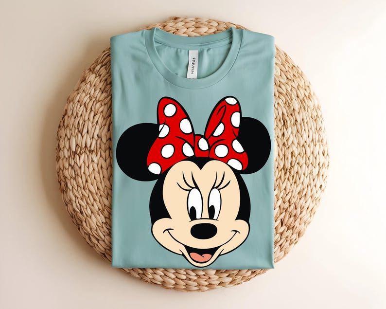 Puede incluir: Una camiseta de cuello redondo en color azul claro con un gr&aacute;fico grande del rostro de Minnie Mouse. El personaje tiene orejas negras, un lazo rojo con lunares blancos y una expresi&oacute;n sonriente. La camiseta tiene una etiqueta blanca en el cuello.