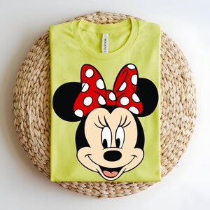 Puede incluir: Una camiseta de cuello redondo color verde lima con un gr&aacute;fico grande del rostro de Minnie Mouse. El personaje tiene orejas negras, cara color crema y un lazo rojo brillante con lunares blancos. La camiseta tiene un corte est&aacute;ndar de manga corta.