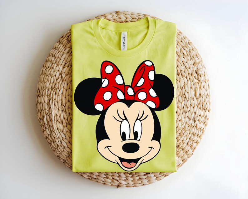 Puede incluir: Una camiseta de cuello redondo color verde lima con un gr&aacute;fico grande del rostro de Minnie Mouse. El personaje tiene orejas negras, cara color crema y un lazo rojo brillante con lunares blancos. La camiseta tiene un corte est&aacute;ndar de manga corta.