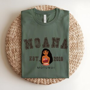 May include: An olive green t-shirt featuring the word "MOANA" in brown varsity lettering. Below the word "MOANA" is the text "EST. 2016" and the word "MOTUNUI". A cartoon image of a woman is in the center.