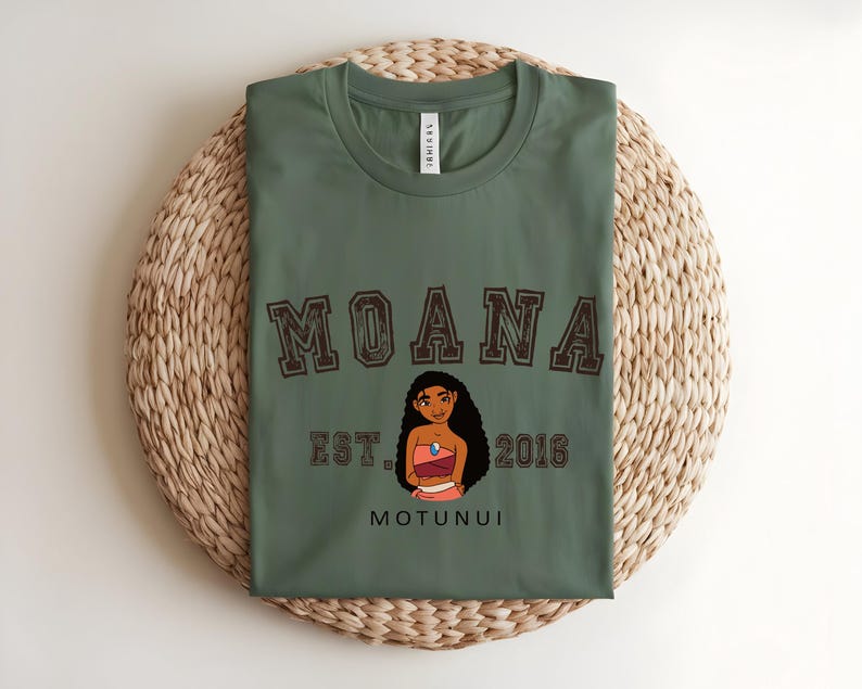 May include: An olive green t-shirt featuring the word "MOANA" in brown varsity lettering. Below the word "MOANA" is the text "EST. 2016" and the word "MOTUNUI". A cartoon image of a woman is in the center.