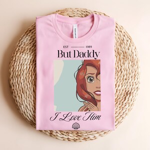 May include: A light pink t-shirt featuring a graphic print of a red-haired character with the text "EST 1989", "But Daddy", "I Love Him", and a small seashell icon. The shirt has a crew neckline and a white brand label visible at the inner collar.