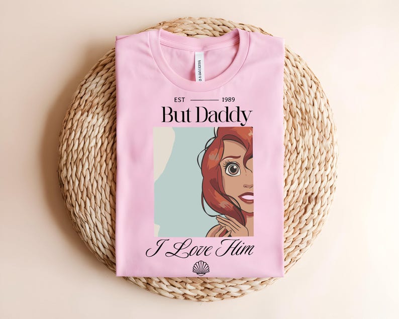 May include: A light pink t-shirt featuring a graphic print of a red-haired character with the text "EST 1989", "But Daddy", "I Love Him", and a small seashell icon. The shirt has a crew neckline and a white brand label visible at the inner collar.