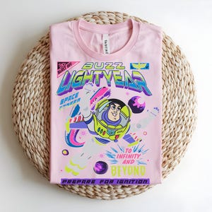 May include: A pale pink t-shirt with a graphic of Buzz Lightyear from Toy Story. The design includes the words "Buzz Lightyear", "Space Ranger", and "To Infinity and Beyond". The shirt is folded on a woven, circular mat.