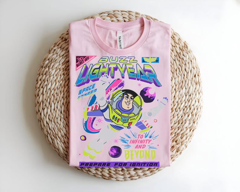May include: A pale pink t-shirt with a graphic of Buzz Lightyear from Toy Story. The design includes the words "Buzz Lightyear", "Space Ranger", and "To Infinity and Beyond". The shirt is folded on a woven, circular mat.