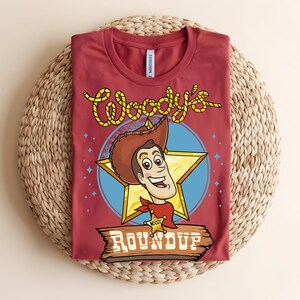 Puede incluir: Una camiseta granate con una imagen de dibujos animados de Woody de Toy Story. El dise&ntilde;o incluye la cara de Woody, una estrella amarilla y las palabras "Woody's Roundup" en una fuente de estilo occidental. La camiseta est&aacute; doblada sobre una estera tejida.