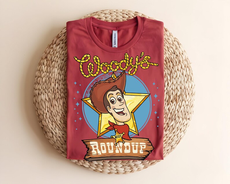 Puede incluir: Una camiseta granate con una imagen de dibujos animados de Woody de Toy Story. El dise&ntilde;o incluye la cara de Woody, una estrella amarilla y las palabras "Woody's Roundup" en una fuente de estilo occidental. La camiseta est&aacute; doblada sobre una estera tejida.
