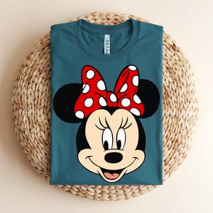 Puede incluir: Una camiseta de cuello redondo color verde azulado con un estampado gr&aacute;fico de Minnie Mouse. El personaje tiene la cabeza negra, orejas grandes y redondas y un lazo rojo brillante con lunares blancos. La etiqueta blanca es visible en el cuello.