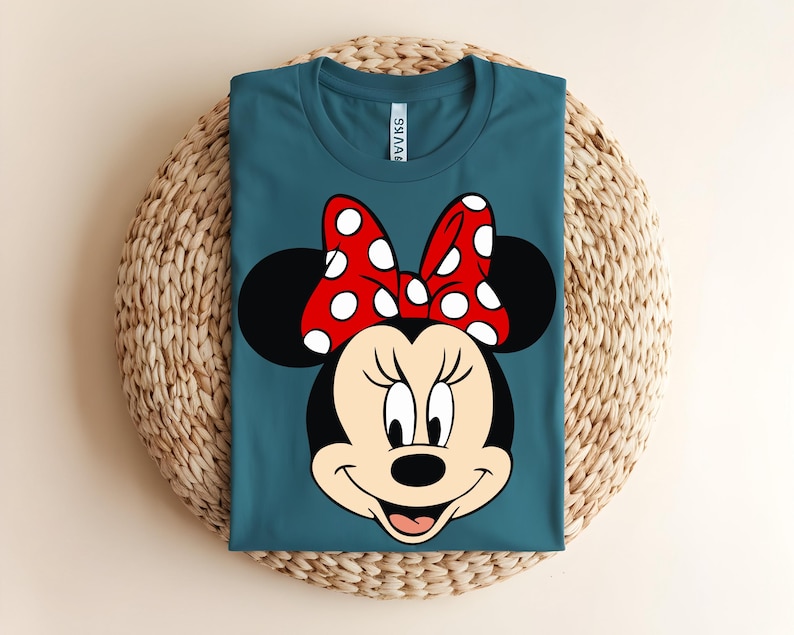 Puede incluir: Una camiseta de cuello redondo color verde azulado con un estampado gr&aacute;fico de Minnie Mouse. El personaje tiene la cabeza negra, orejas grandes y redondas y un lazo rojo brillante con lunares blancos. La etiqueta blanca es visible en el cuello.