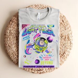 May include: A pale grey t-shirt with a vibrant graphic of Buzz Lightyear. The design includes the words "Buzz Lightyear," "To Infinity and Beyond," and "Prepare for Ignition." The shirt is folded on a woven mat.