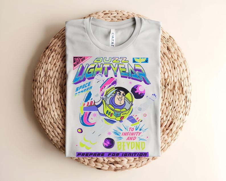 May include: A pale grey t-shirt with a vibrant graphic of Buzz Lightyear. The design includes the words "Buzz Lightyear," "To Infinity and Beyond," and "Prepare for Ignition." The shirt is folded on a woven mat.