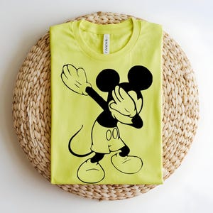 K&ouml;nnte beinhalten: Ein leuchtend neongelbes T-Shirt mit Rundhalsausschnitt und einem gro&szlig;en schwarzen Grafikdruck von Mickey Mouse in einer Dab-Pose. Das T-Shirt hat ein klassisches Kurzarm-Design mit einem sichtbaren wei&szlig;en Markenetikett im Kragenbereich.
