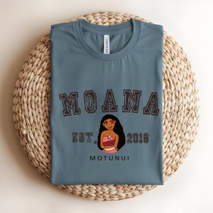 May include: A blue t-shirt with the word "MOANA" in a brown, collegiate-style font. Below the word "MOANA" is a cartoon illustration of a woman, the text "EST. 2016", and the word "MOTUNUI". The shirt is folded on a woven, circular mat.