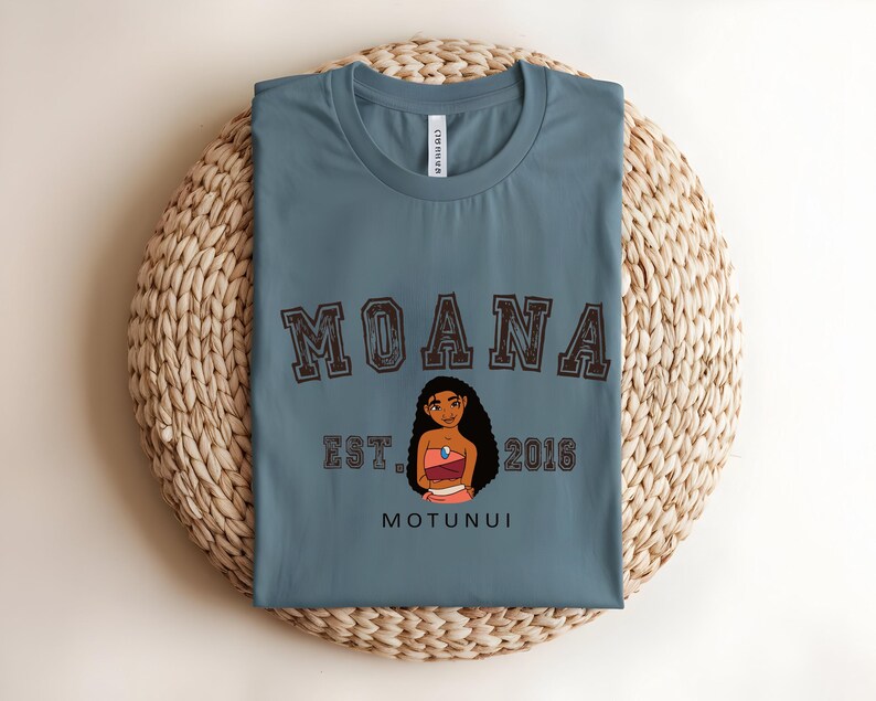 May include: A blue t-shirt with the word "MOANA" in a brown, collegiate-style font. Below the word "MOANA" is a cartoon illustration of a woman, the text "EST. 2016", and the word "MOTUNUI". The shirt is folded on a woven, circular mat.