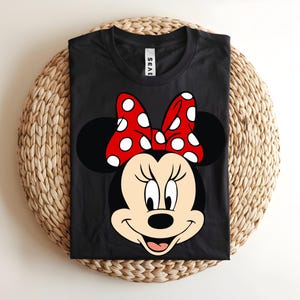 Puede incluir: Una camiseta negra de cuello redondo con un gran estampado gr&aacute;fico de Minnie Mouse en la parte delantera. El personaje tiene la cara color crema, orejas negras y un lazo rojo brillante con lunares blancos. La camiseta tiene una etiqueta blanca en el cuello con texto negro.