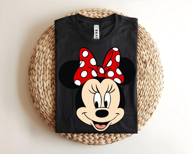 Puede incluir: Una camiseta negra de cuello redondo con un gran estampado gr&aacute;fico de Minnie Mouse en la parte delantera. El personaje tiene la cara color crema, orejas negras y un lazo rojo brillante con lunares blancos. La camiseta tiene una etiqueta blanca en el cuello con texto negro.