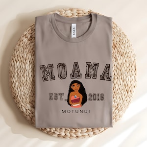 May include: A grey t-shirt with the word "MOANA" in a brown collegiate font above a cartoon character illustration. Below the illustration are the words "EST. 2016" and "MOTUNUI". The shirt is folded on a woven mat.