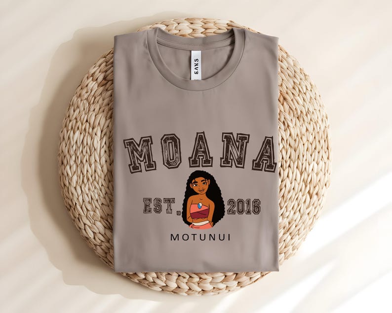 May include: A grey t-shirt with the word "MOANA" in a brown collegiate font above a cartoon character illustration. Below the illustration are the words "EST. 2016" and "MOTUNUI". The shirt is folded on a woven mat.