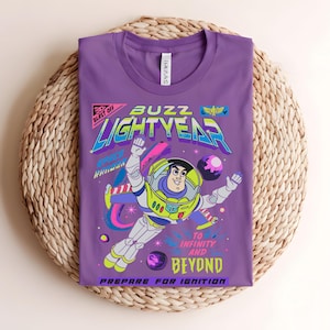 May include: A purple t-shirt with a graphic of Buzz Lightyear from Toy Story. The design includes the words "Buzz Lightyear" and "To Infinity and Beyond." The graphic is in bright colours with a retro style.