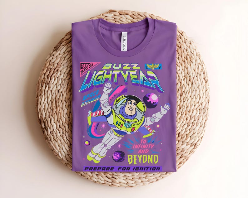 May include: A purple t-shirt with a graphic of Buzz Lightyear from Toy Story. The design includes the words "Buzz Lightyear" and "To Infinity and Beyond." The graphic is in bright colours with a retro style.