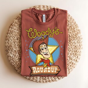 Puede incluir: Una camiseta de color &oacute;xido con una ilustraci&oacute;n de dibujos animados de Woody de Toy Story. El dise&ntilde;o incluye la cara de Woody, una estrella amarilla y el texto "Woody's Roundup" en una fuente de estilo occidental. La camiseta se muestra sobre una estera tejida.