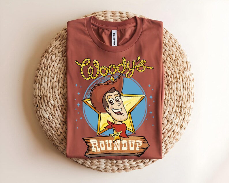 Puede incluir: Una camiseta de color &oacute;xido con una ilustraci&oacute;n de dibujos animados de Woody de Toy Story. El dise&ntilde;o incluye la cara de Woody, una estrella amarilla y el texto "Woody's Roundup" en una fuente de estilo occidental. La camiseta se muestra sobre una estera tejida.