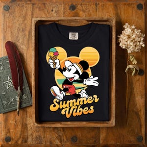 以下が含まれることがあります： ミッキーマウスがアイスクリームを持っている黒いTシャツ。背景は黄色、オレンジ、緑の夕焼けのデザイン。「Summer Vibes」の文字は黄色で書かれています。木製のトレイに入っています。