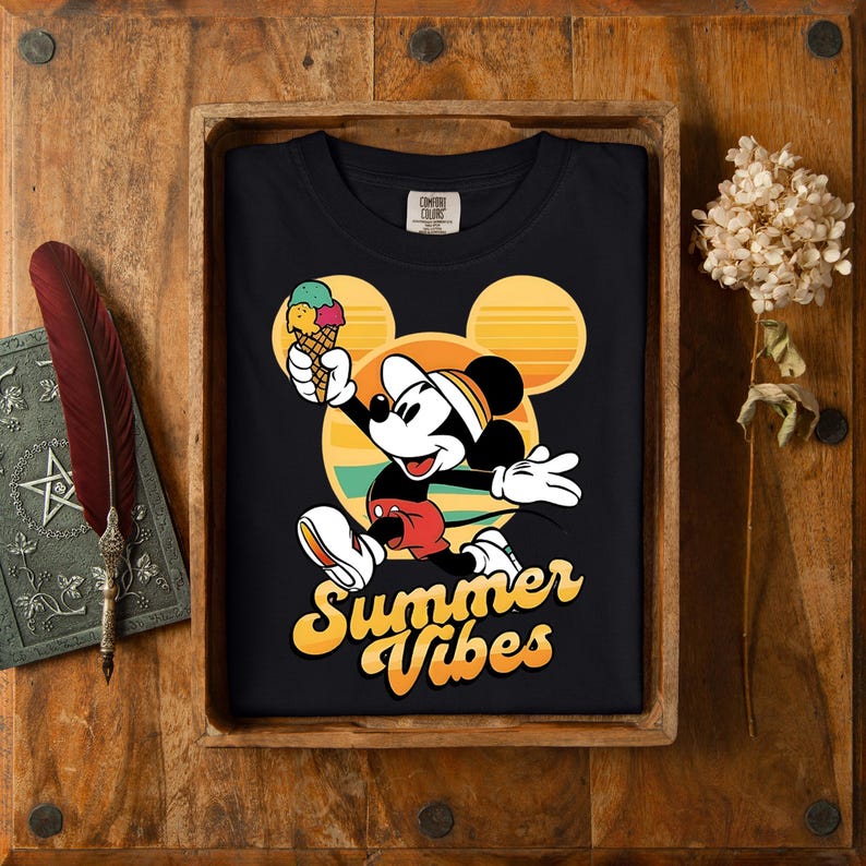 以下が含まれることがあります： ミッキーマウスがアイスクリームを持っている黒いTシャツ。背景は黄色、オレンジ、緑の夕焼けのデザイン。「Summer Vibes」の文字は黄色で書かれています。木製のトレイに入っています。