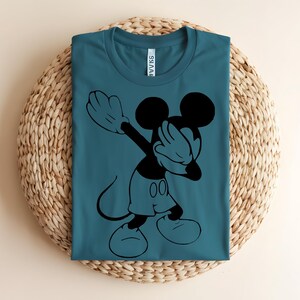 K&ouml;nnte beinhalten: Ein blaugr&uuml;nes T-Shirt mit Rundhalsausschnitt und einem schwarzen Grafikdruck von Mickey Mouse, der eine Dab-Pose ausf&uuml;hrt. Das Shirt hat ein klassisches Kurzarm-Design mit einem sichtbaren wei&szlig;en Markenetikett im inneren Nackenbereich.