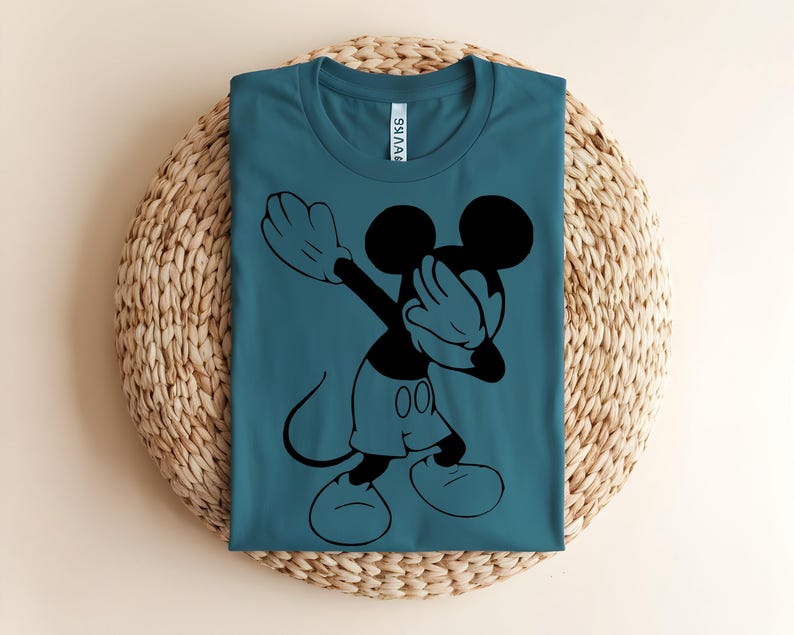 K&ouml;nnte beinhalten: Ein blaugr&uuml;nes T-Shirt mit Rundhalsausschnitt und einem schwarzen Grafikdruck von Mickey Mouse, der eine Dab-Pose ausf&uuml;hrt. Das Shirt hat ein klassisches Kurzarm-Design mit einem sichtbaren wei&szlig;en Markenetikett im inneren Nackenbereich.