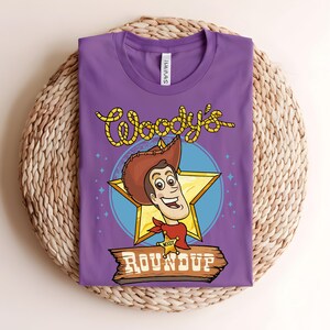 Puede incluir: Una camiseta morada con una ilustraci&oacute;n de dibujos animados de Woody de Toy Story. El dise&ntilde;o incluye la cara de Woody, una estrella y el texto "Woody's Roundup" en amarillo y marr&oacute;n. La camiseta est&aacute; doblada sobre una superficie tejida.
