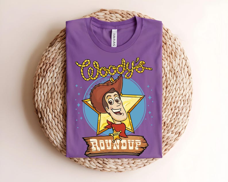 Puede incluir: Una camiseta morada con una ilustraci&oacute;n de dibujos animados de Woody de Toy Story. El dise&ntilde;o incluye la cara de Woody, una estrella y el texto "Woody's Roundup" en amarillo y marr&oacute;n. La camiseta est&aacute; doblada sobre una superficie tejida.