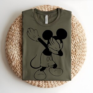 K&ouml;nnte beinhalten: Ein olivgr&uuml;nes T-Shirt mit Rundhalsausschnitt und einem schwarzen Grafikdruck der Figur Mickey Mouse, die eine Dab-Pose ausf&uuml;hrt. Das Shirt hat ein klassisches Kurzarm-Design mit einem sichtbaren Markenetikett im inneren Nackenbereich.