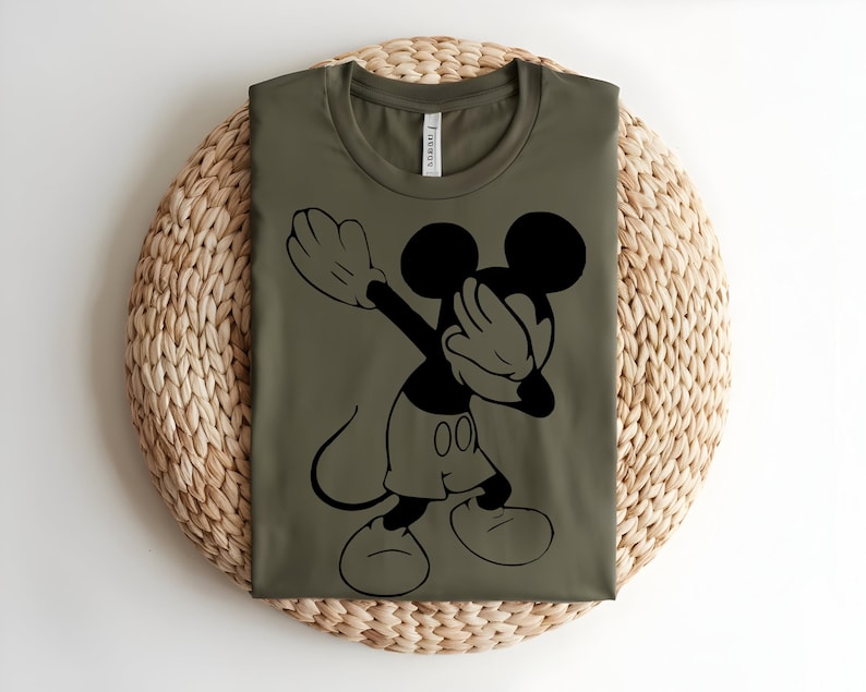 K&ouml;nnte beinhalten: Ein olivgr&uuml;nes T-Shirt mit Rundhalsausschnitt und einem schwarzen Grafikdruck der Figur Mickey Mouse, die eine Dab-Pose ausf&uuml;hrt. Das Shirt hat ein klassisches Kurzarm-Design mit einem sichtbaren Markenetikett im inneren Nackenbereich.
