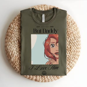 May include: An olive green t-shirt featuring a graphic of a red-haired character with wide eyes. The text reads "EST 1989 But Daddy I Love Him" in black lettering, with a small seashell icon centered at the bottom of the graphic design.