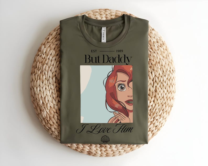 May include: An olive green t-shirt featuring a graphic of a red-haired character with wide eyes. The text reads "EST 1989 But Daddy I Love Him" in black lettering, with a small seashell icon centered at the bottom of the graphic design.