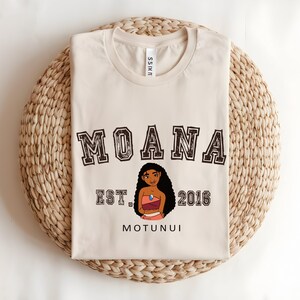 May include: A cream-coloured t-shirt with the word "MOANA" in a collegiate font. Below is an illustration of a cartoon character, with "EST. 2016" and "MOTUNUI" beneath. The shirt is folded on a woven, circular mat.