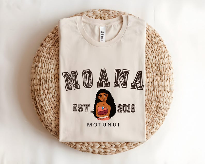 May include: A cream-coloured t-shirt with the word "MOANA" in a collegiate font. Below is an illustration of a cartoon character, with "EST. 2016" and "MOTUNUI" beneath. The shirt is folded on a woven, circular mat.