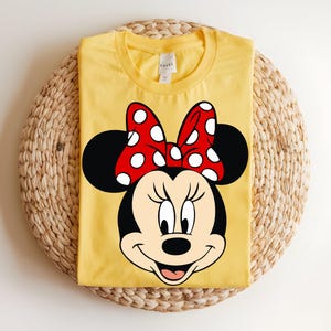 Puede incluir: Una camiseta de color amarillo brillante con cuello redondo que presenta un gran estampado gr&aacute;fico de Minnie Mouse en la parte delantera. El personaje aparece con sus caracter&iacute;sticas orejas negras y un lazo rojo vivo con lunares blancos.