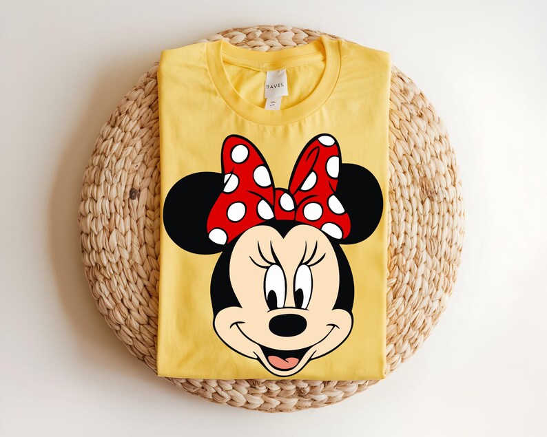 Puede incluir: Una camiseta de color amarillo brillante con cuello redondo que presenta un gran estampado gr&aacute;fico de Minnie Mouse en la parte delantera. El personaje aparece con sus caracter&iacute;sticas orejas negras y un lazo rojo vivo con lunares blancos.