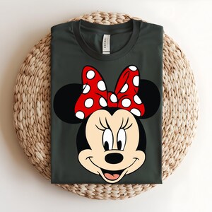Puede incluir: Una camiseta de cuello redondo gris oscuro con un gr&aacute;fico de Minnie Mouse. El personaje tiene la cabeza negra, orejas grandes y redondas y un lazo rojo brillante con lunares blancos. El dise&ntilde;o est&aacute; centrado en el pecho de la camiseta.